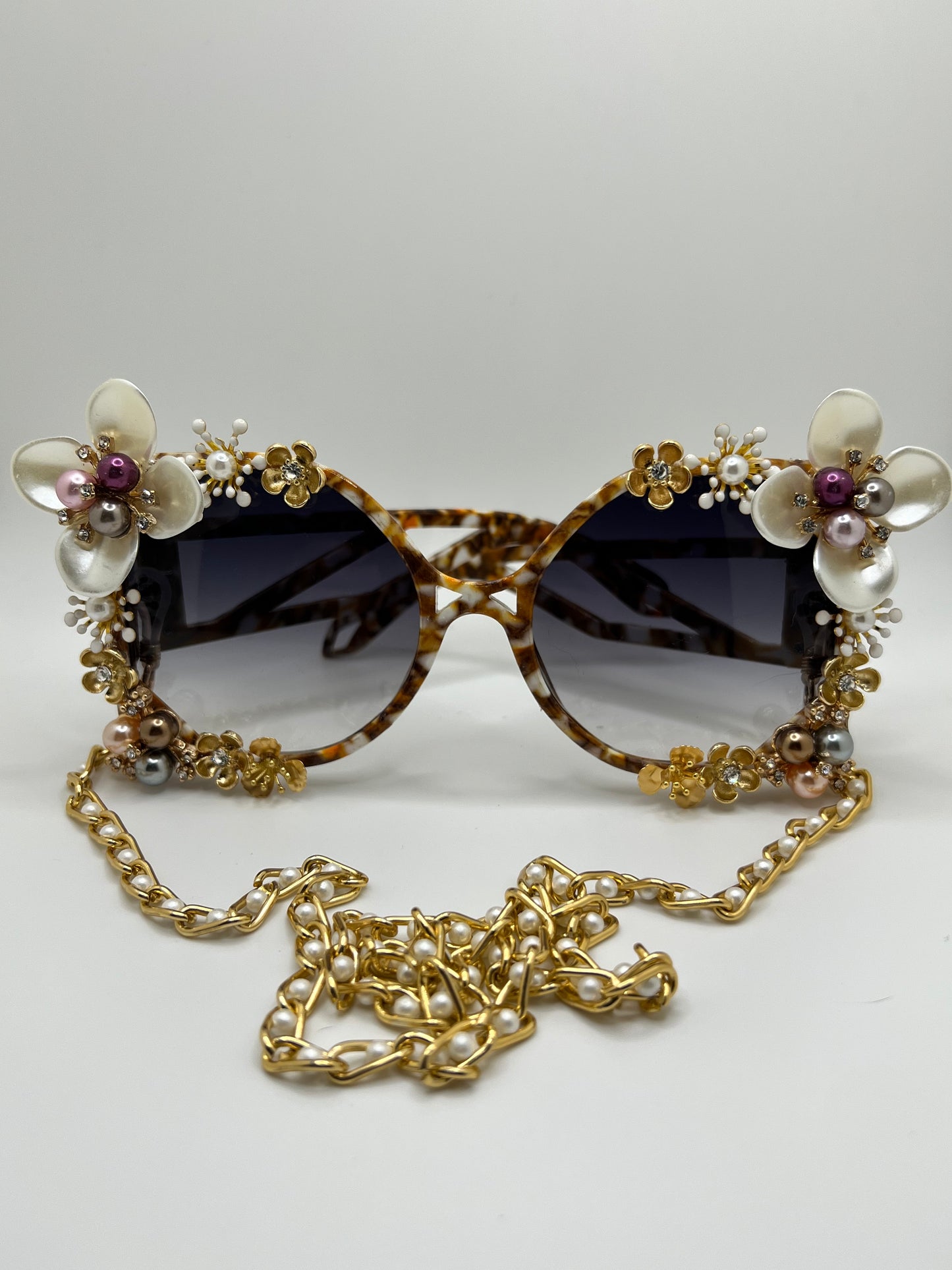 These square-sided, oversized sunglasses are covered in pearl flowers In white, pink, and deep maroon and are accompanied by an adjustable and removable eyewear chain.
