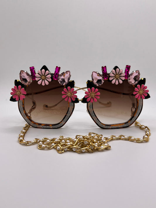 Hues of bright pink jewels and flowers surround these polygon, glittering sunglasses, and are accompanied by an adjustable and removable eyewear chain.